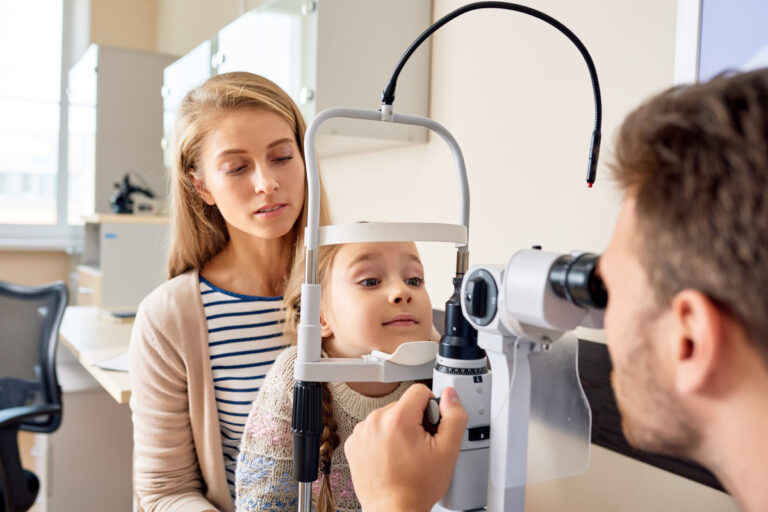 Portrait of cute little girl siting on mothers lap looking at slit lamp machine during medical check up in eye clinic