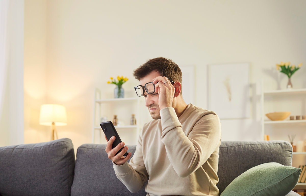 Portrait of a young man sitting on sofa at home holding eyeglasses and trying to read message on smartphone having farsightedness eye disease. Presbyopia and vision problems concept.