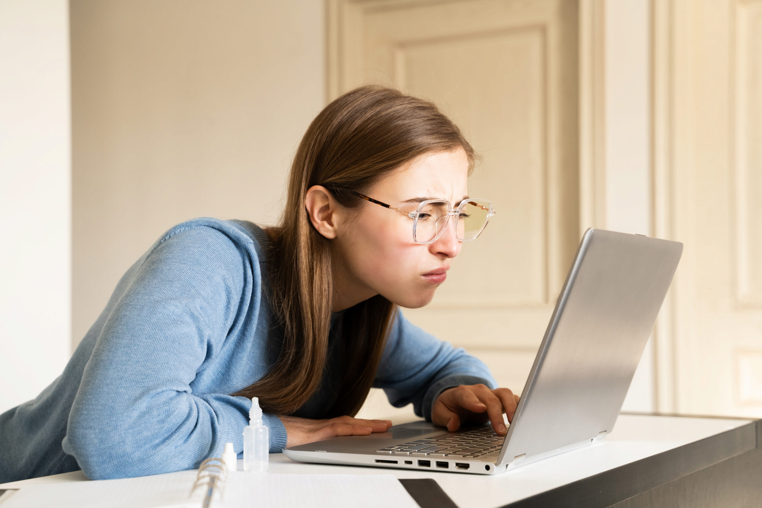 Woman with poor eyesight wearing eyeglasses looking too close at laptop screen at desk during work or study at home. Female squinting eyes. Ophthalmology bad blurry vision digital eye strain symptoms.