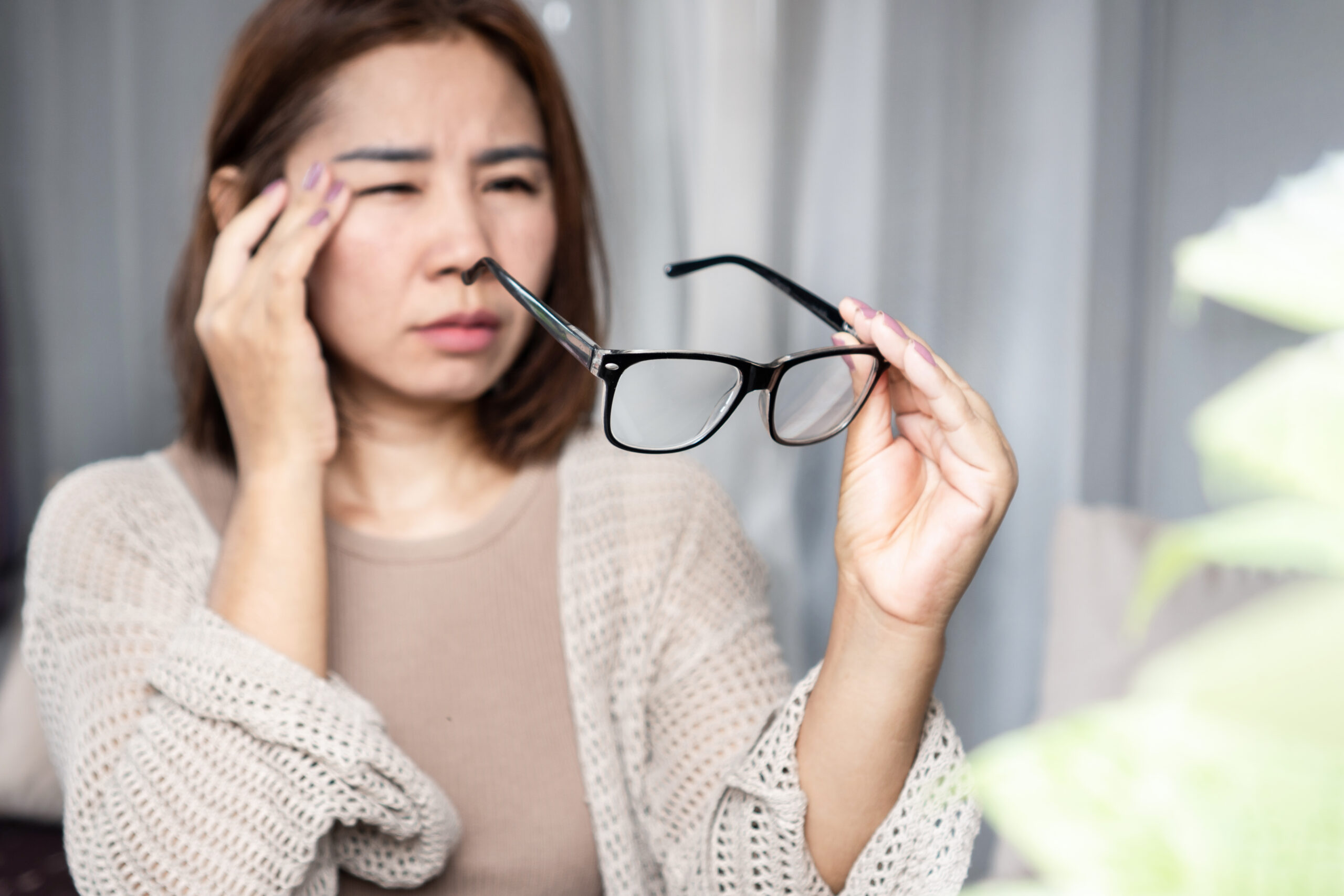 Asian woman feeling a headache, dizziness, and blurry vision while holding her eyeglasses, showing signs of eye fatigue and vision problems such as nearsightedness or farsightedness