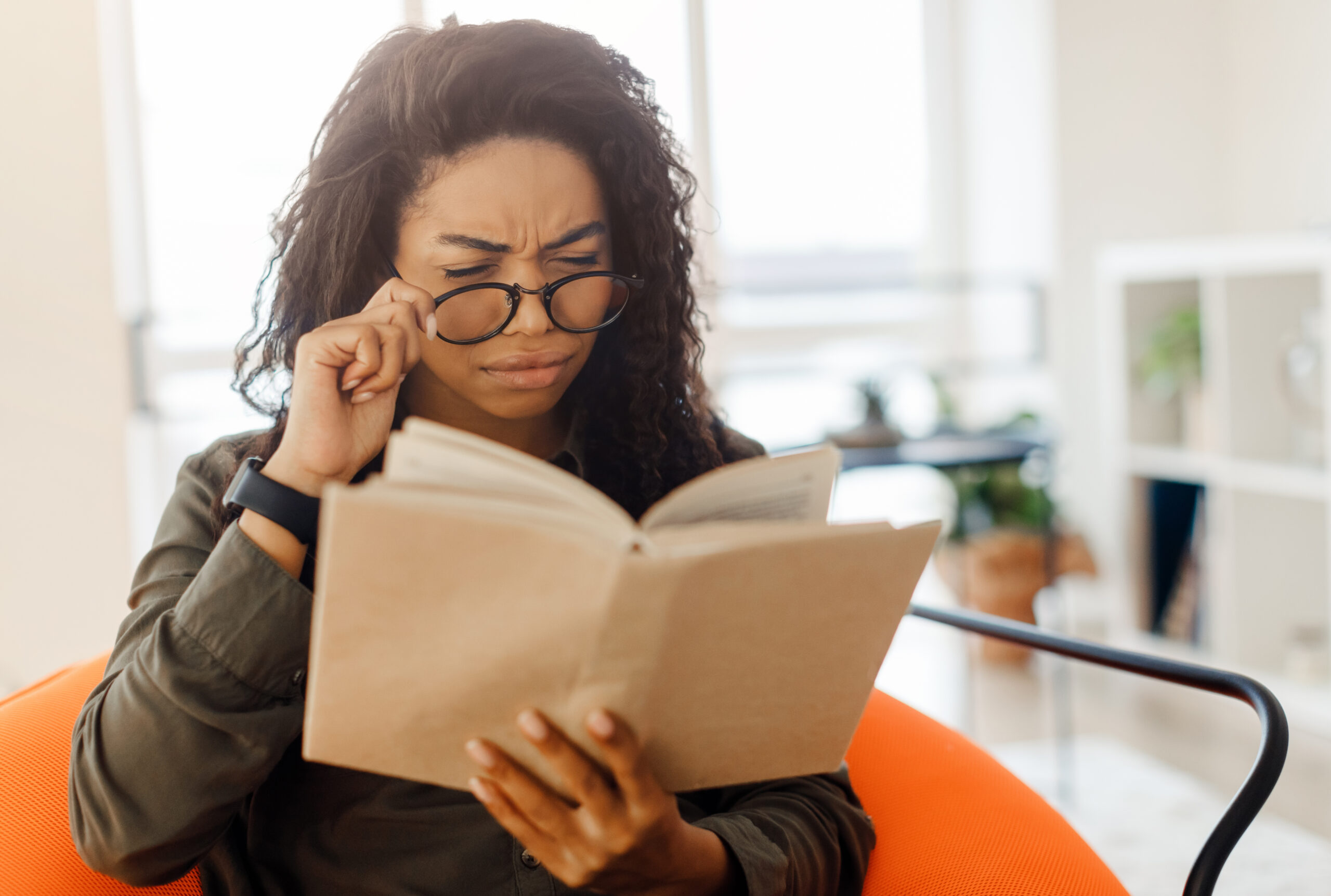 Focused confused young black lady trying to read paper book, squinting to see more clearly, wearing glasses, having difficulties seeing text because of vision problems, sitting on pouf in living room