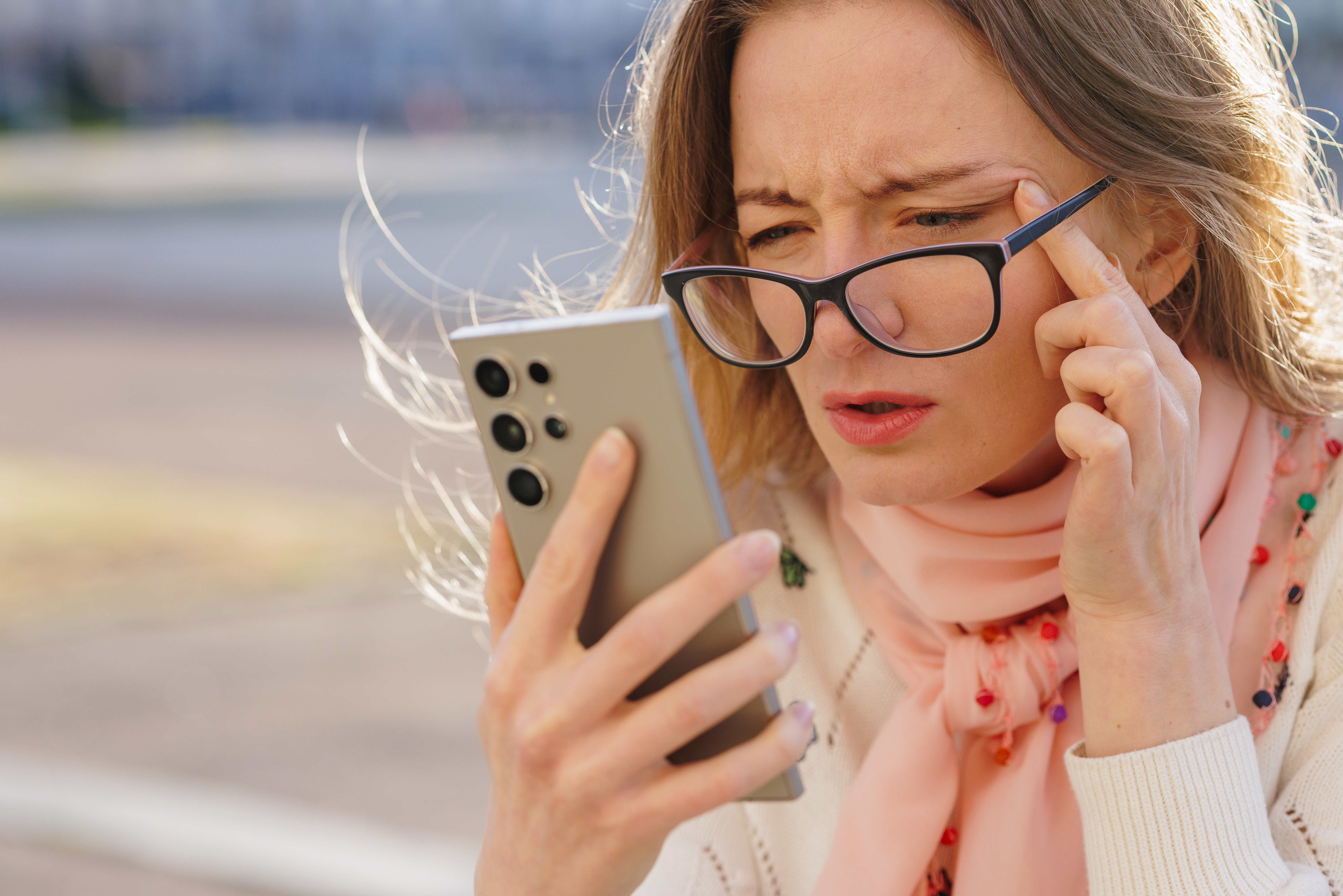 Woman struggling to read phone screen outdoors, squinting through glasses, showing symptoms of poor eyesight and digital fatigue.