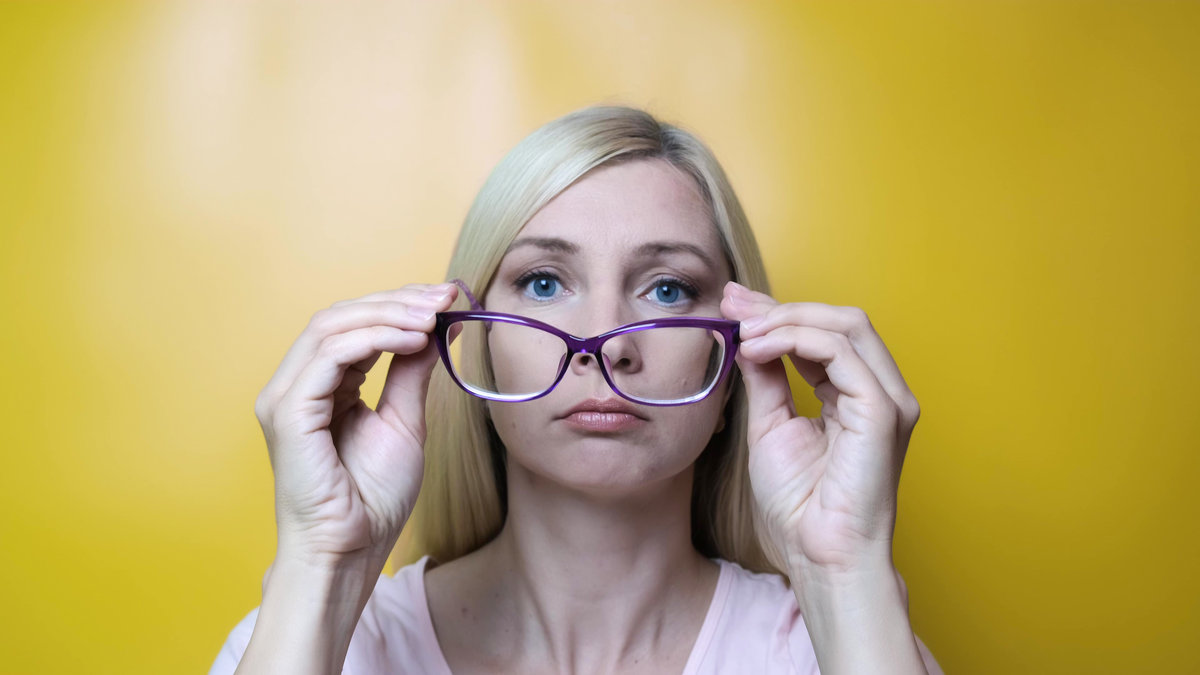 A middle age blond woman putting on stylish fashionable glasses and smiling to camera against yellow background, myopia, astigmatism and ophthalmologic diseases comcept.