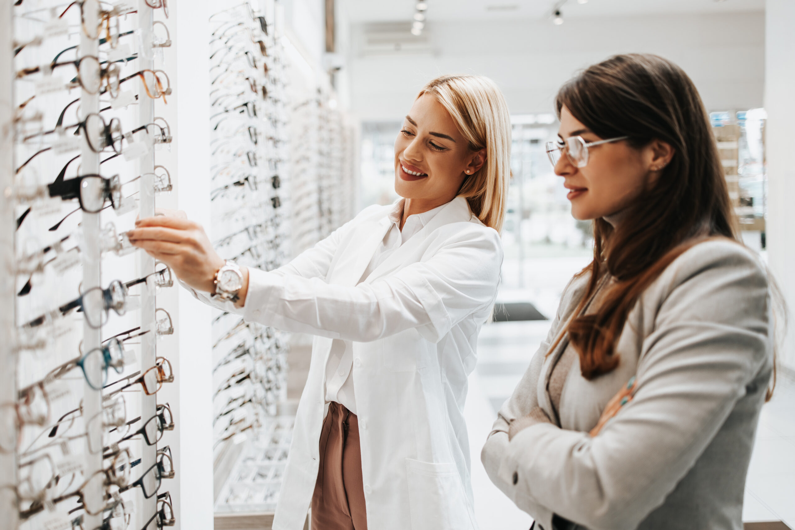 Beautiful and fashionable woman choosing eyeglasses frame in modern optical store. Female seller specialist helps her to make right decision.