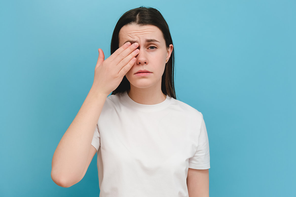 Young girl suffering from eyes pain and feeling something in eye, posing on blue wall. Cause of pain include contact lens problem, conjunctivitis, foreign object, dry eye syndrome or allergy