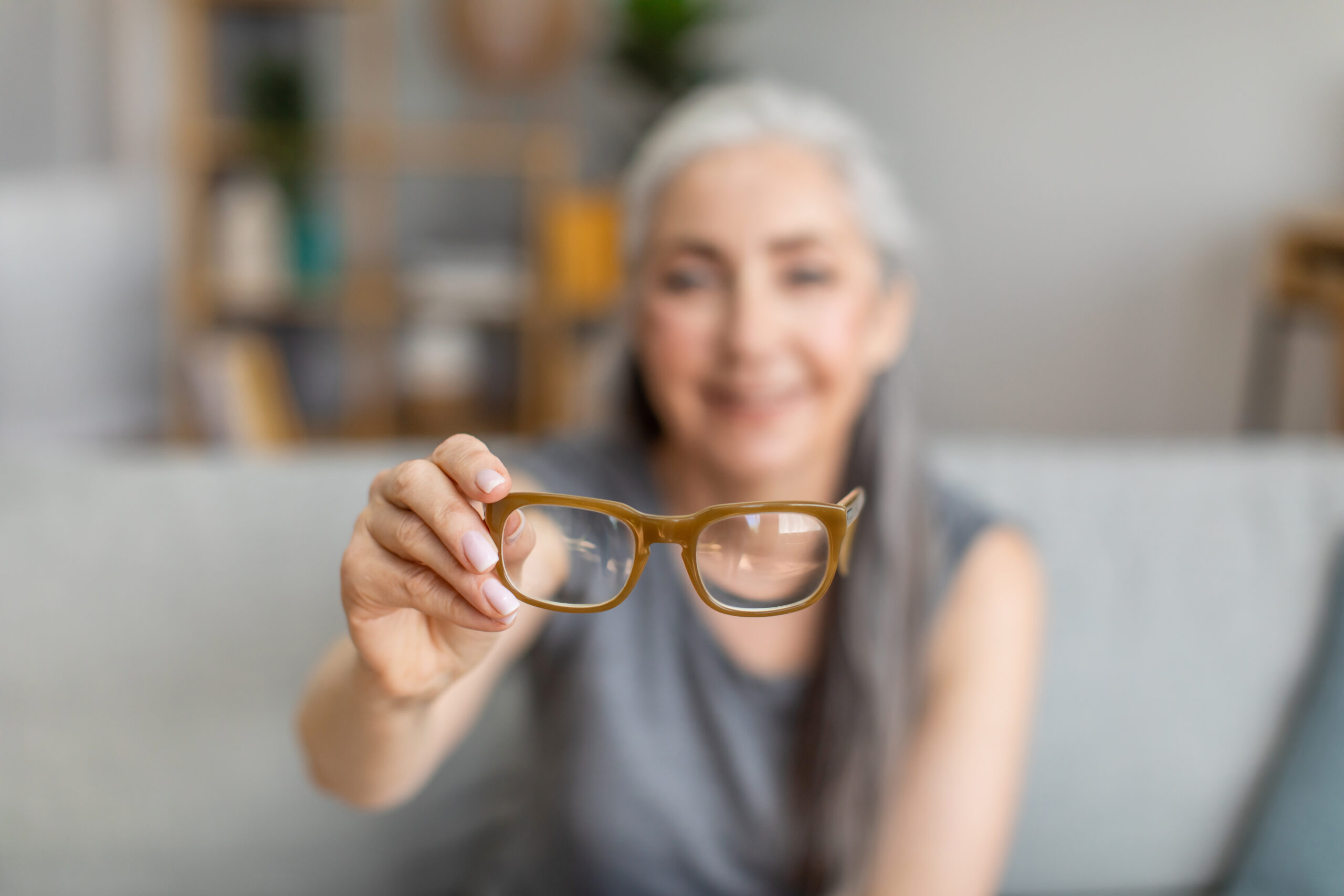 Cheerful caucasian elderly gray-haired female holding glasses in living room interior, selective focus, blurry. Health care, poor eyesight, age-related problems, myopia and advertising, free space