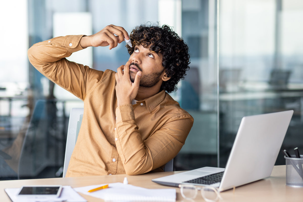 A young businessman works for a long time at a laptop inside the office, the man drips drops into his eyes, eye pain. An Indian man sits with a laptop at the workplace.