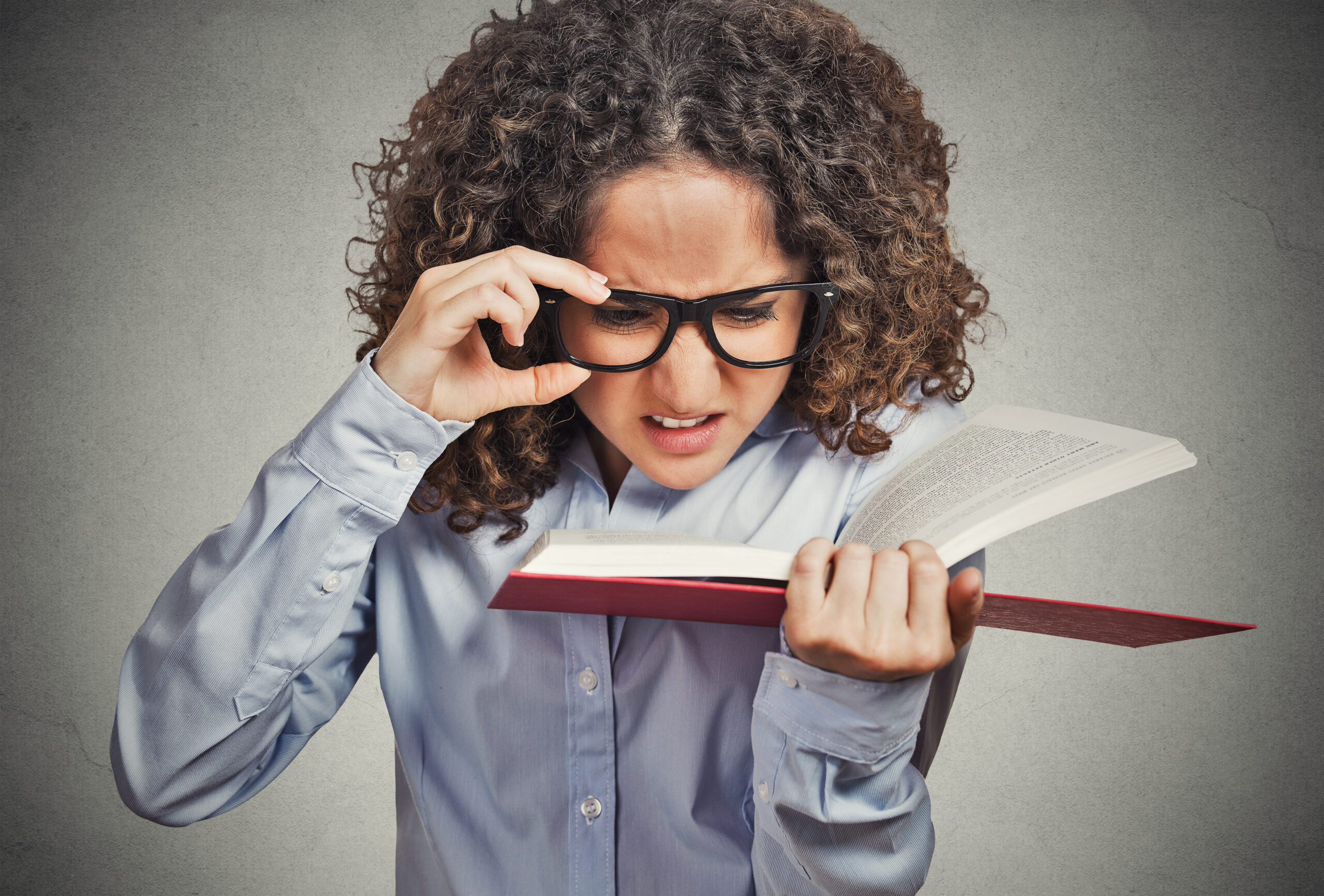 Closeup portrait young woman with eye glasses trying to read book, having difficulties seeing text, bad vision sight problem isolated grey background. Face expression reaction health eyesight issues