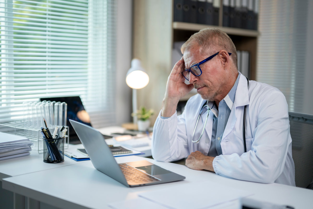 Stressed mature male doctor in white coat sitting at desk in office looking concerned at laptop screen
