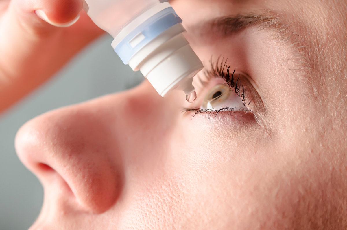 Closeup view of young woman applying eye drop, artificial tears. Cute girl suffering from an ophthalmological allergic reaction to pollen. Girl applying artificial tears sitting on a sofa in the living room at home. Close up view of young woman applying eye drop to left eye, artificial tears, antibiotics, antihistamine.