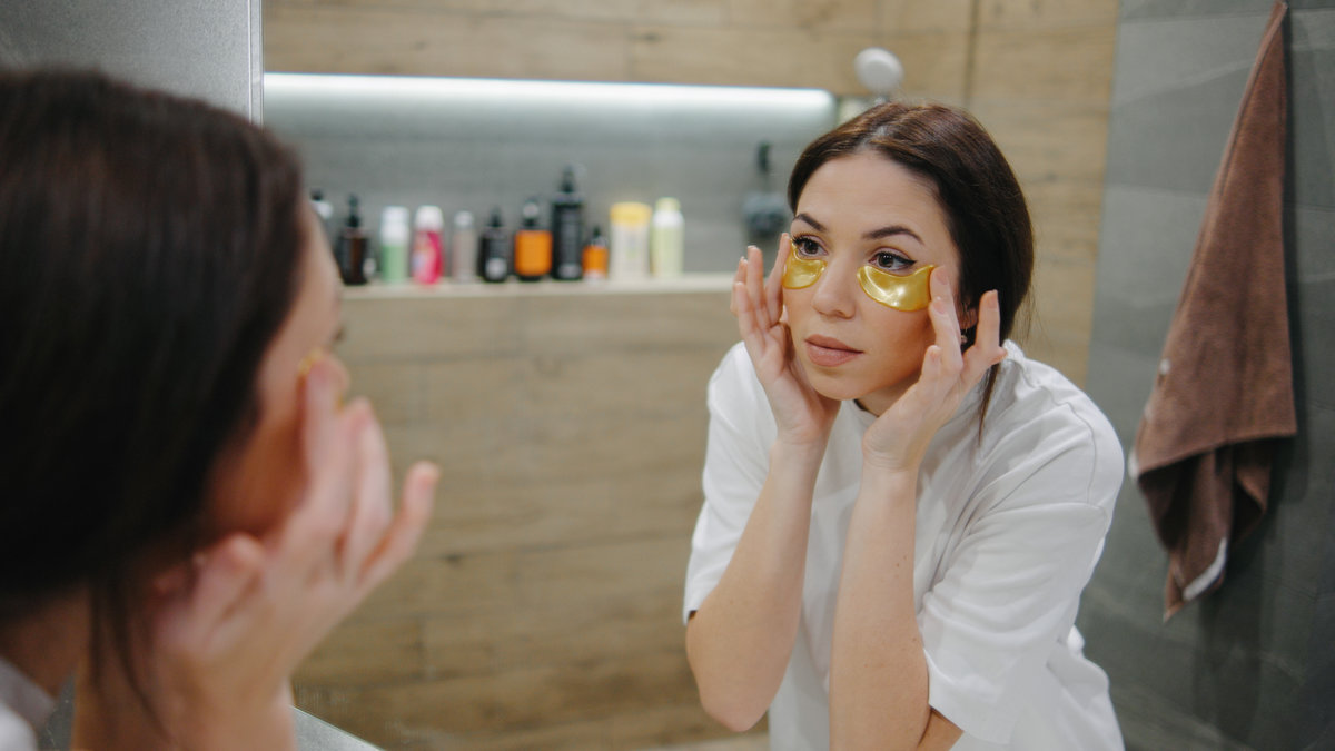 A woman stands in a stylish bathroom, applying yellow eye patches to her under-eyes while checking her reflection in the mirror.