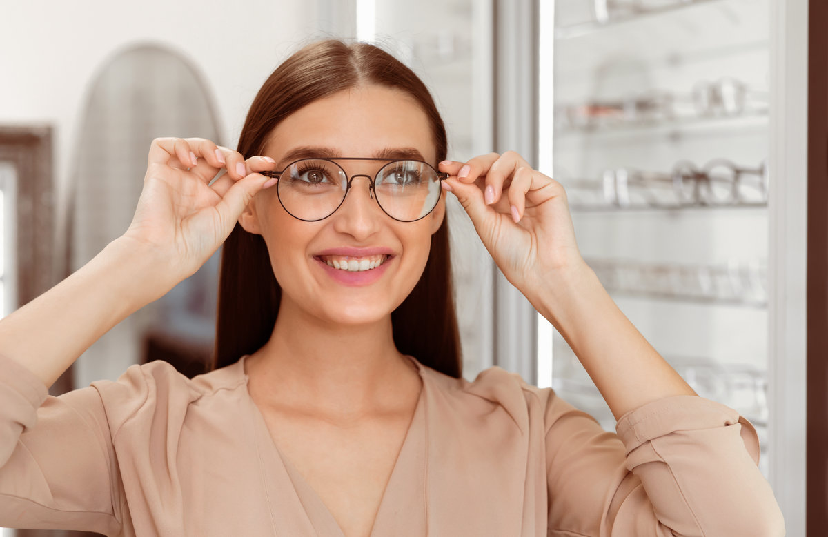 A young woman smiles while adjusting a pair of fashionable glasses in an eyewear store. Sunlight illuminates her happy expression as she explores her options.