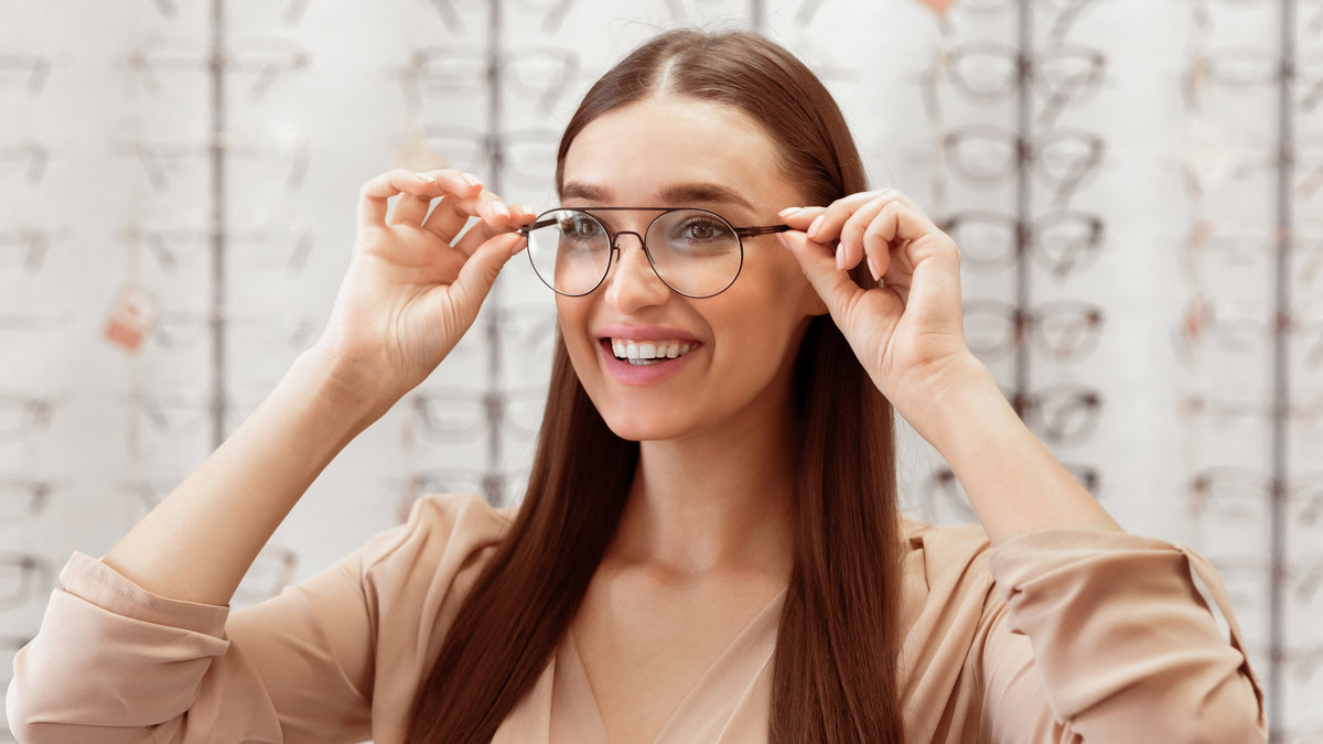 A young woman is smiling while adjusting her eyeglasses in an optical store filled with various frames. She appears excited about her new look and options available.