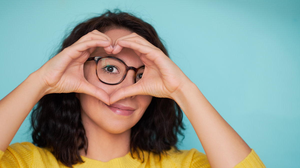 woman in glasses on a turquoise background makes a heart shape with her hands, a symbol of love.