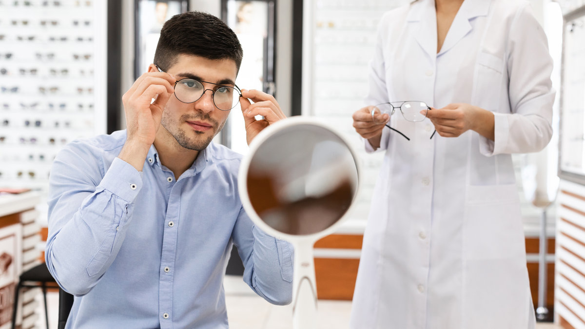 Vision Correction Concept. Female optometrist helping male client to choose eyeglasses for computer, holding specs frame, guy trying on spectacles, sitting at desk and looking at mirror