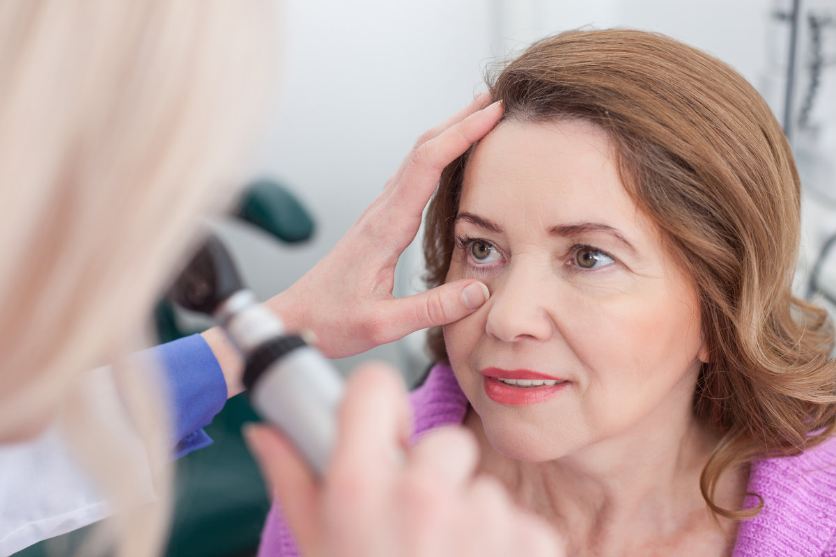 Skillful oculist is examining female eyes in clinic. She is holding a special tool and directing laser on female eye. The woman is sitting and smiling