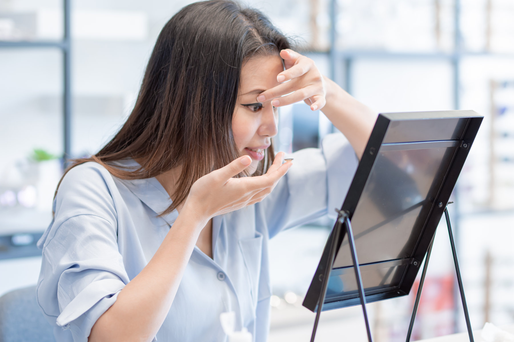 Asian beautiful woman trying to put contact lenses into her eyes and looking to the mirror. Lifestyle and Optic Concept.