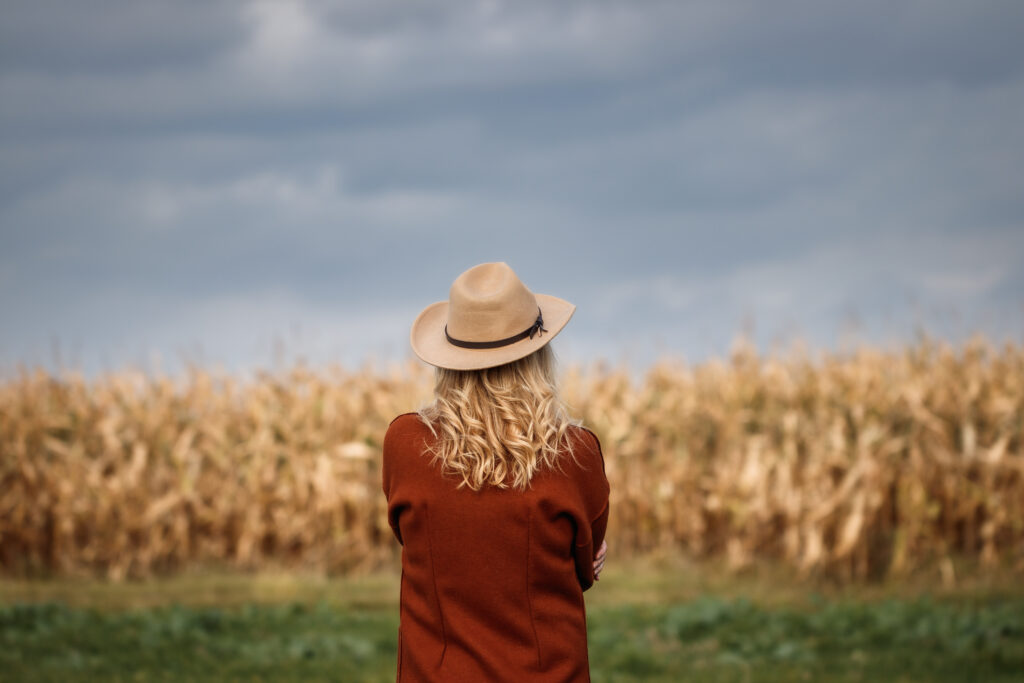 Woman with red coat and cowboy hat looks at cloudy sky over corn field
