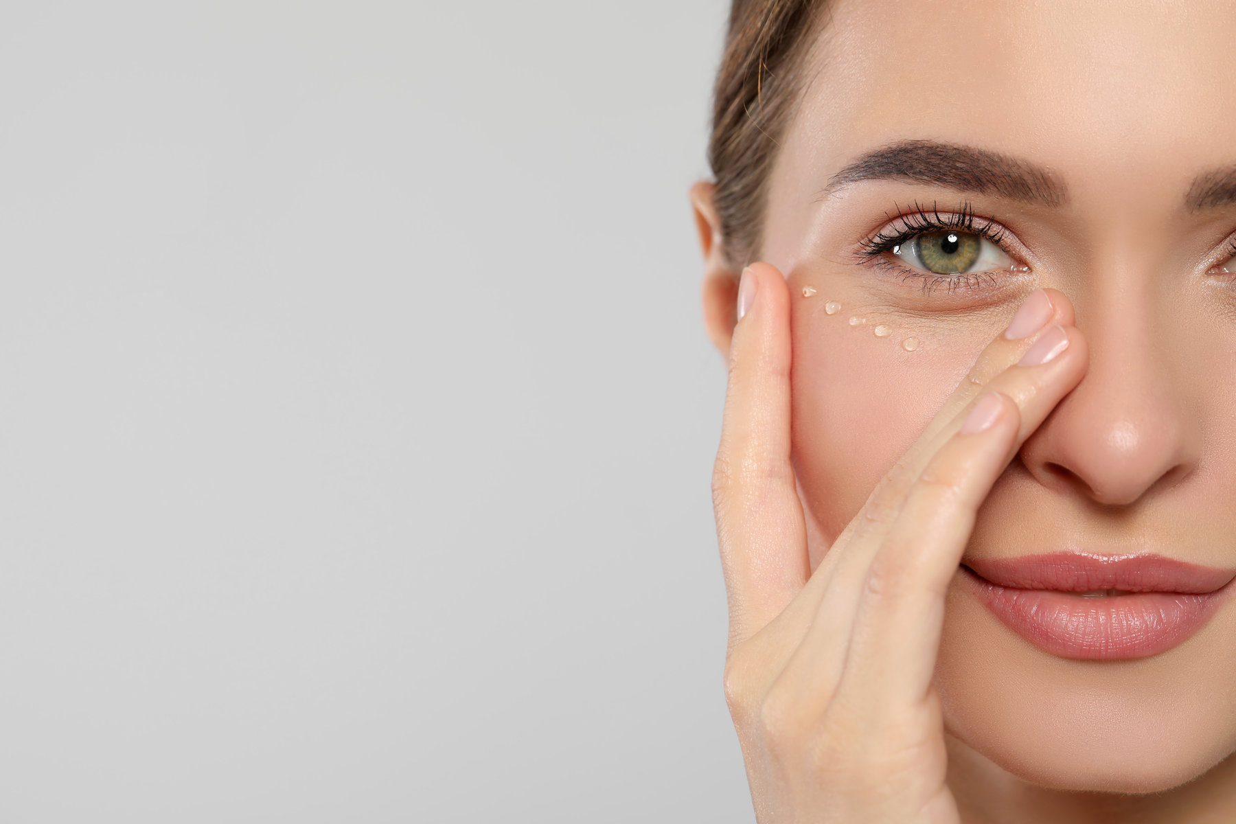 Young woman with cream around eye on white background, closeup. Space for text